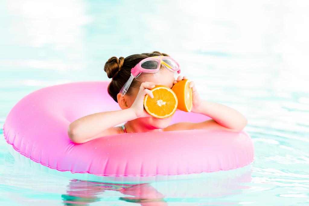 Kind hält halbe Orangen in Augennähe beim Schwimmen mit aufblasbarem Ring im Schwimmbad - Foto, Bild