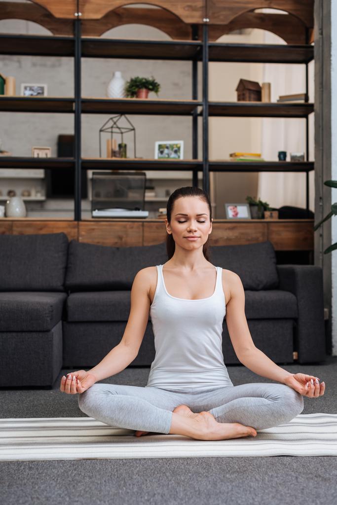 kaunis nuori nainen istuu lootuksessa poseeraa ja meditoi kotona olohuoneessa
 - Valokuva, kuva