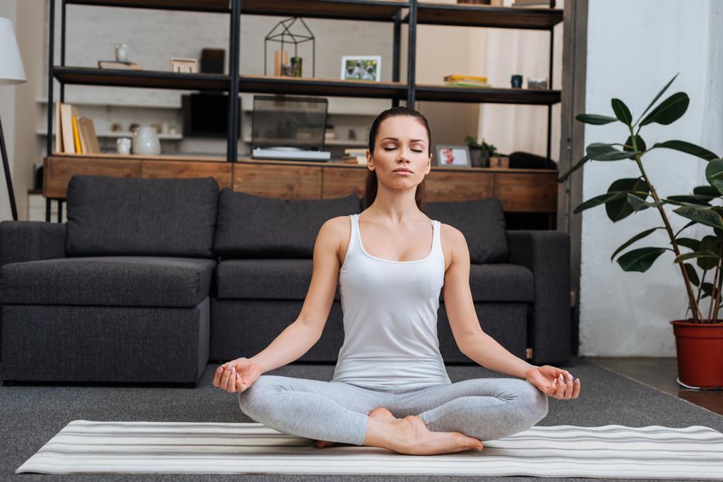 beautiful young woman sitting in lotus pose and meditating at home in living room - Photo, Image