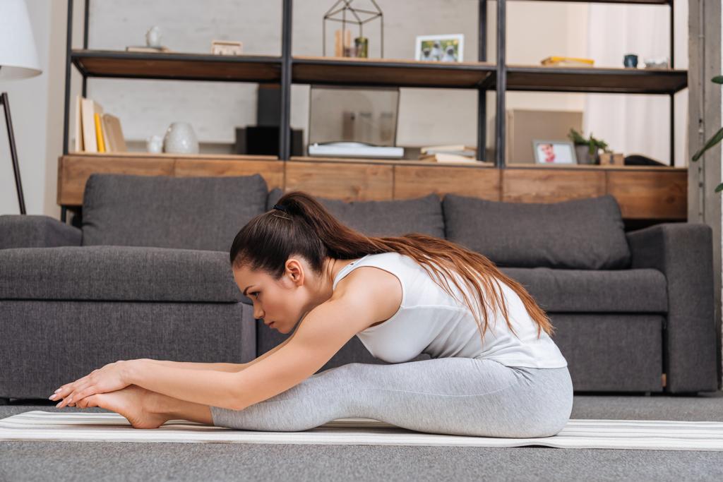 Frau übt sitzend vorwärts klappbare Pose auf Fitnessmatte zu Hause im Wohnzimmer - Foto, Bild