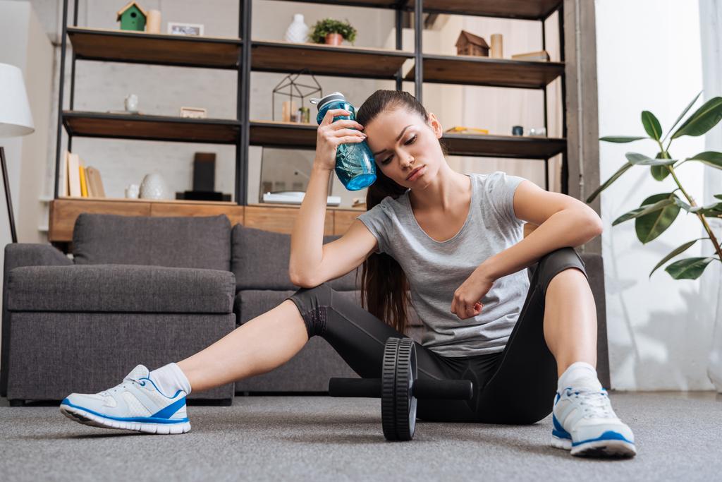 tired sportswoman with sport bottle after training with ab wheel at home - Photo, Image
