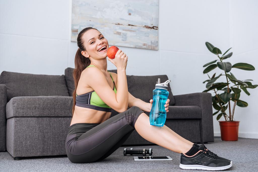 usmívající se sportovkyně doma drží láhev s vodou a jablko - Fotografie, Obrázek