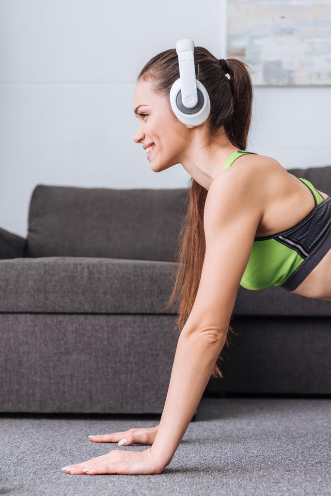 красива усміхнена спортсменка в навушниках робить дошку вправи вдома
 - Фото, зображення