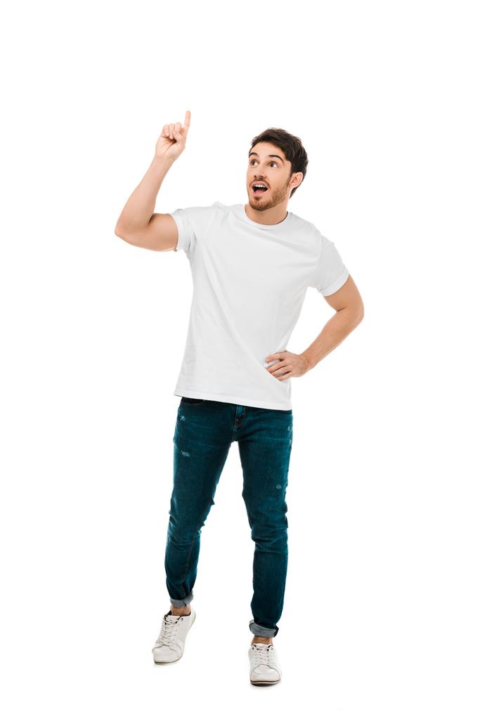full length view of young man standing with hand on waist and pointing up with finger isolated on white - Photo, Image