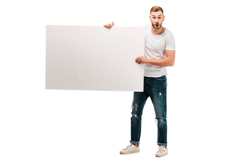 σοκαρισμένος νεαρός άνδρας κρατώντας πλακάτ κενό και βλέπουν τα φωτογραφικών μηχανών που απομονώνονται σε λευκό - Φωτογραφία, εικόνα