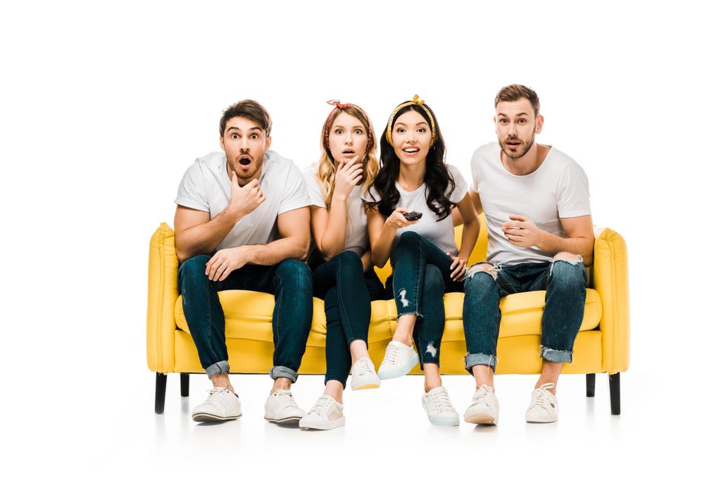 shocked young friends sitting on sofa with remote controller and looking at camera isolated on white - Photo, Image