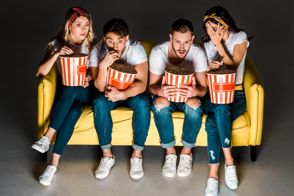 vista de ángulo alto de jóvenes amigos asustados sosteniendo palomitas de maíz y viendo la televisión mientras se sientan juntos en el sofá en gris
  - Foto, Imagen