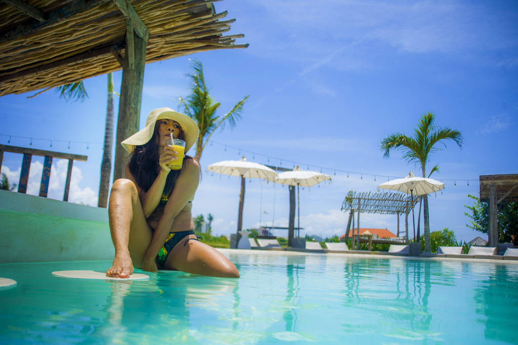 Młoda sexy i atrakcyjna egzotycznych azjatyckich Kobieta w bikini i kapelusz lato picie soku w paradise resort basen bar uśmiechający się zadowolony korzystających luksusowe wakacje wycieczka w tropikalnej wyspie - Zdjęcie, obraz