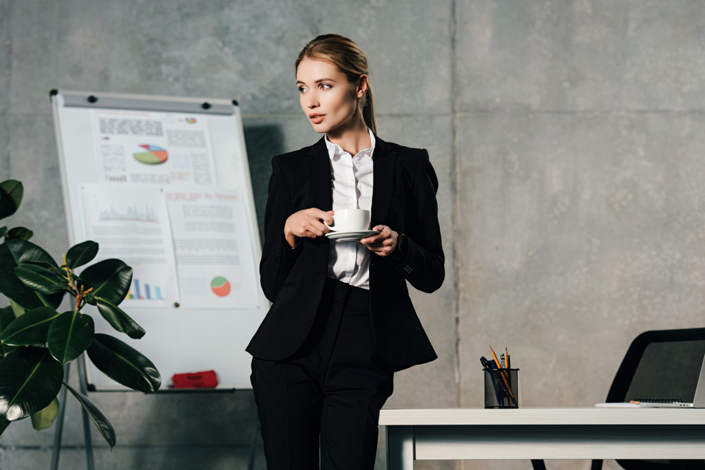 junge Geschäftsfrau trinkt Kaffee, während sie im Büro am Arbeitsplatz steht - Foto, Bild