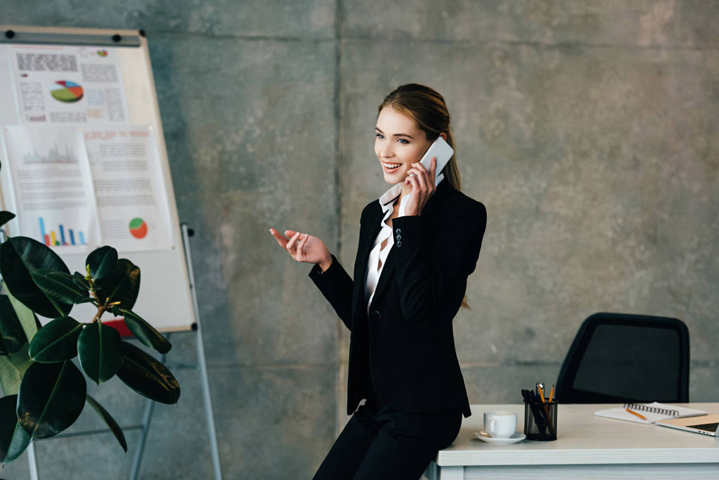 усміхнена бізнес-леді говорить на смартфоні, стоячи за робочим столом
  - Фото, зображення