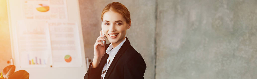sonriente mujer de negocios hablando en el teléfono inteligente y mirando a la cámara en la oficina
 - Foto, Imagen
