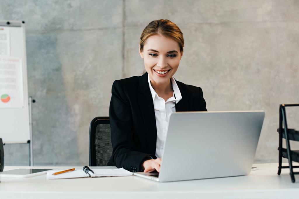 усміхнена бізнес-леді сидить на робочому місці і використовує ноутбук
  - Фото, зображення