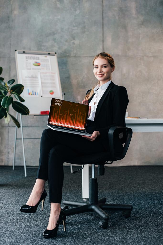χαμογελαστός νεαρός επιχειρηματίας κάθεται σε καρέκλα και να κατέχουν φορητό υπολογιστή με το διαδικτυακό εμπόριο τιμές στην οθόνη - Φωτογραφία, εικόνα