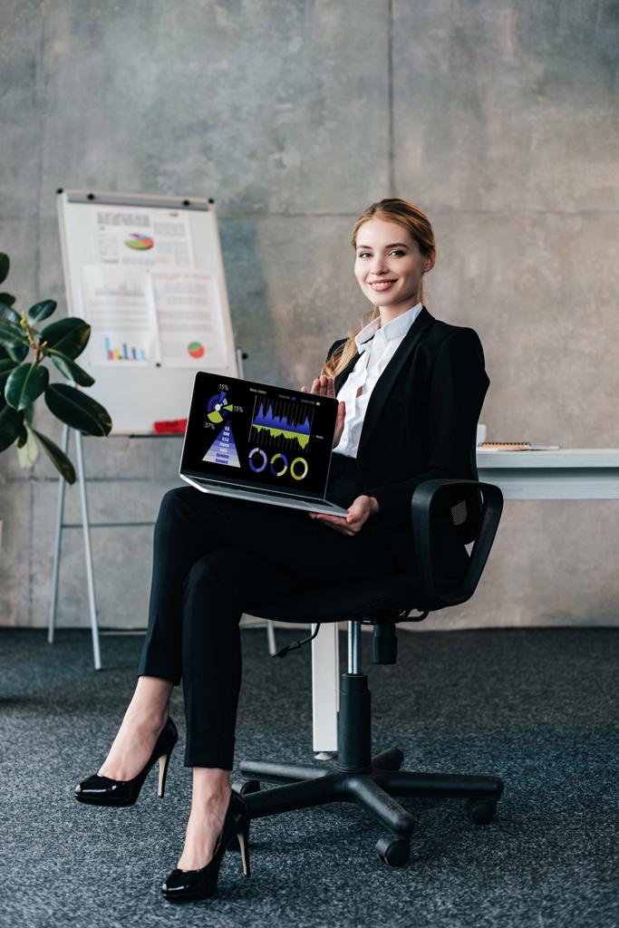 Glimlachende zakenvrouw zittend op een stoel en holding laptop met commerciële diagrammen op scherm - Foto, afbeelding