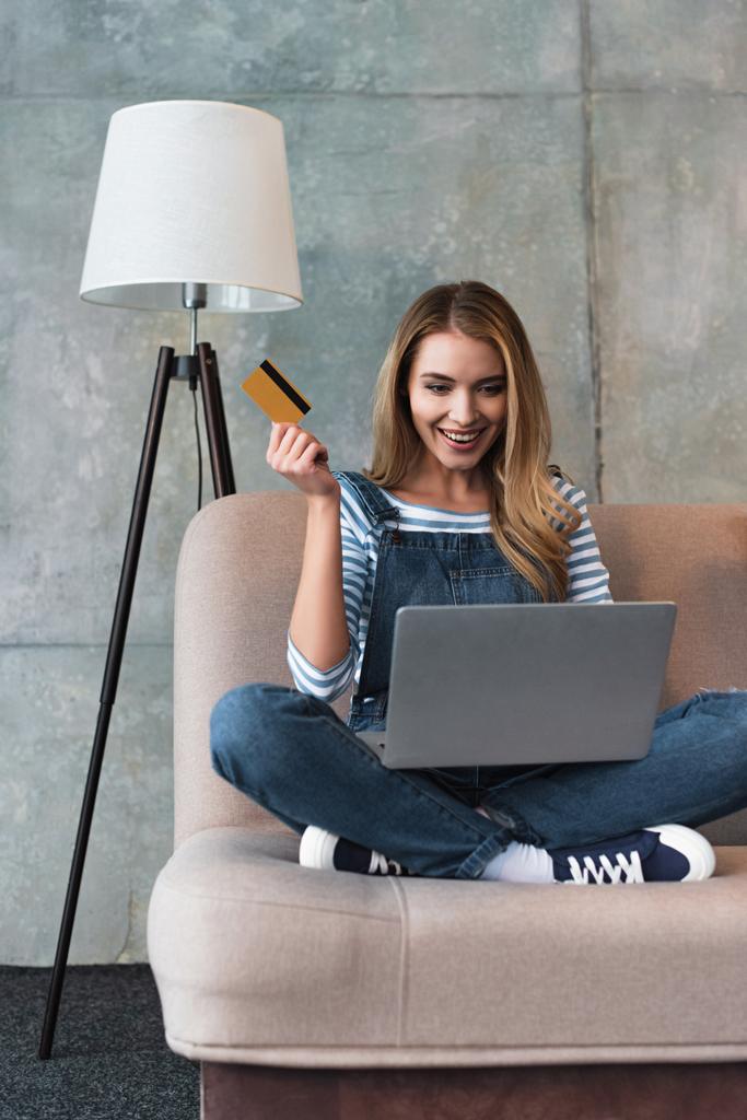 happy beautiful woman holding credit card in hand and sitting on sofa with laptop  - Photo, Image