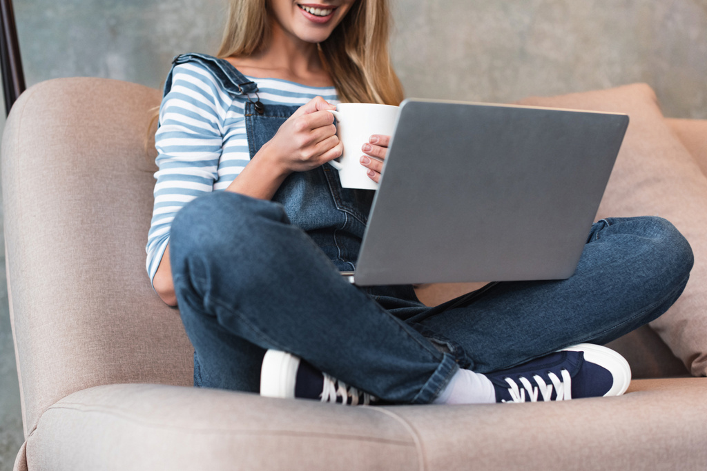 schöne Frau lächelt und sitzt mit Tasse und Laptop auf dem Sofa - Foto, Bild