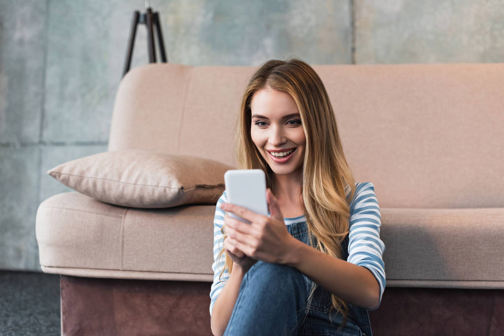 Młoda kobieta uśmiechając się, za pomocą smartfona i siedzi w pobliżu różowy sofa - Zdjęcie, obraz