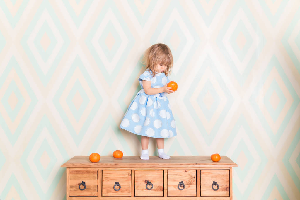 Enfant en bas âge fille en robe bleu clair debout sur la commode en bois et tenant mandarine orange fraîche dans la main et en le regardant dans la surprise étirant ses lèvres sur fond de papier peint losange
 - Photo, image