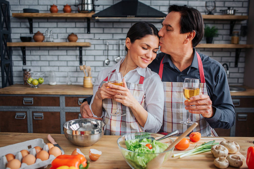 Lovely picture of man sitting with woman together at table in kitchen. He kiss her forehead and hold glass on wine in hand. woman lean to him and enjoy. - Photo, Image