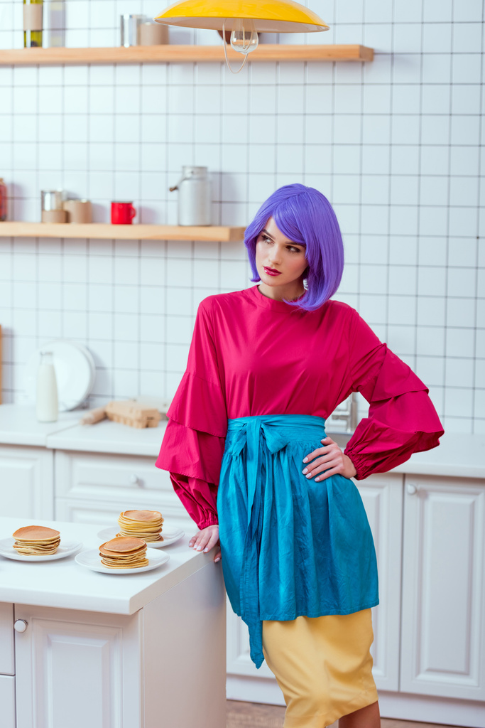 valikoiva painopiste kotiäiti violetti hiukset ja värikkäitä vaatteita poseeraa lähellä keittiö counter pannukakkuja
 - Valokuva, kuva