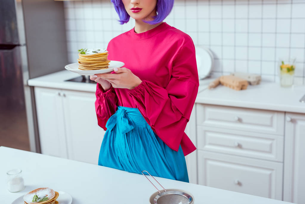 частичный вид домохозяйки с фиолетовыми волосами и красочной одеждой, держащей тарелку с блинами на кухне
 - Фото, изображение