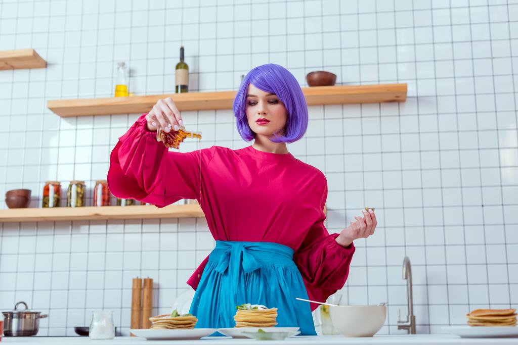 gerichte huisvrouw met paarse haren en kleurrijke kleding gieten van siroop op pannenkoeken in keuken - Foto, afbeelding