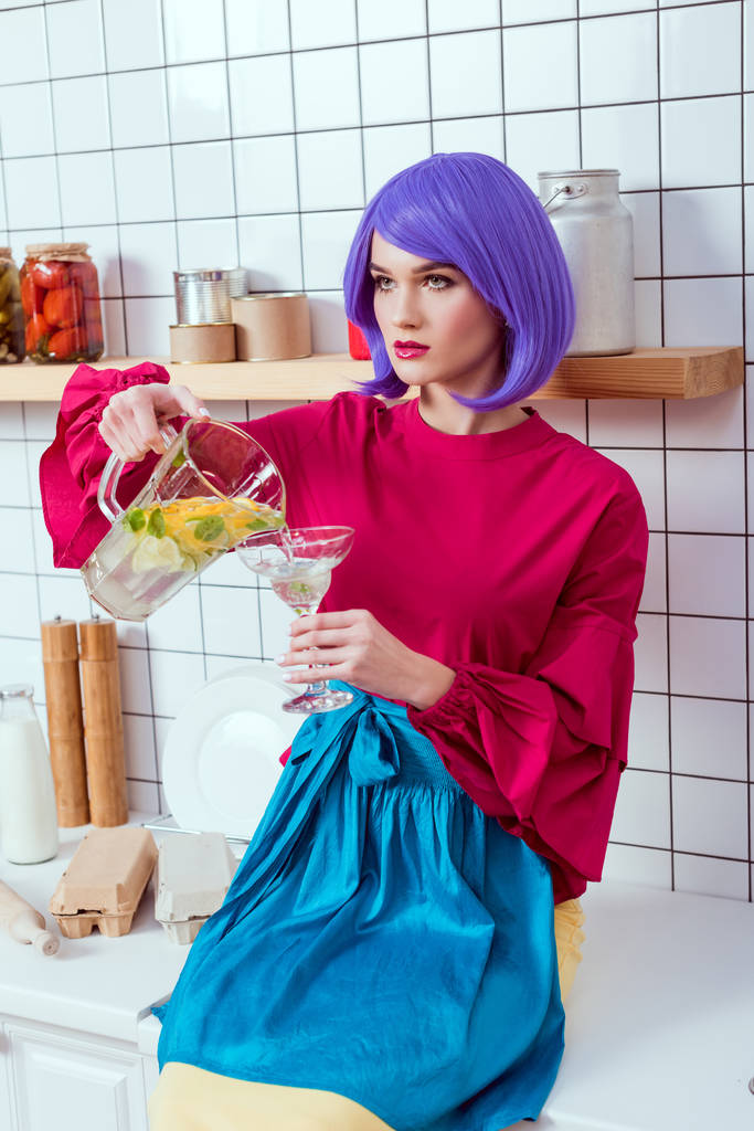 casalinga con capelli viola e vestiti colorati seduta sul bancone della cucina e versando limonata in vetro
 - Foto, immagini