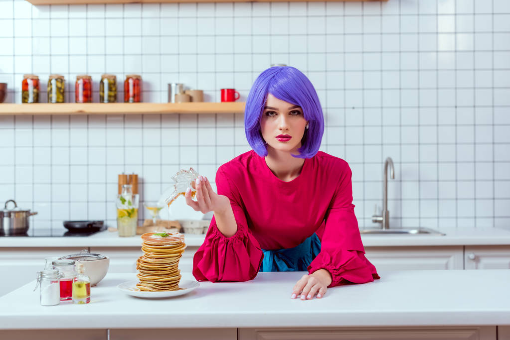 όμορφη νοικοκυρά με μοβ μαλλιά και πολύχρωμα ρούχα έκχυση σιρόπι για τηγανίτες, ενώ κοιτάζοντας τα φωτογραφικών μηχανών στην κουζίνα - Φωτογραφία, εικόνα