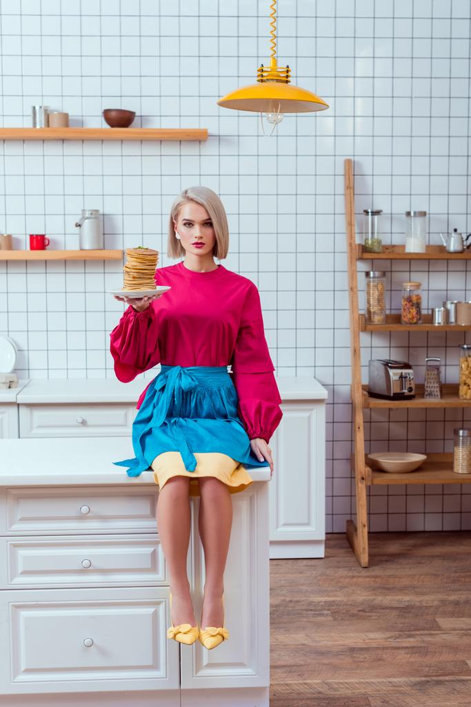 μοντέρνα γυναίκα σε πολύχρωμα ρούχα κρατώντας την πλάκα των τηγανίτες και κοιτάζοντας την κάμερα στην κουζίνα - Φωτογραφία, εικόνα
