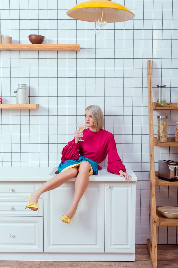 hermosa mujer de moda sentada en el mostrador de la cocina con copa de cóctel y mirando hacia otro lado
 - Foto, Imagen