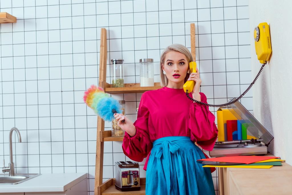 surprised housewife in colorful clothes holding dusting brush and talking on retro telephone in kitchen - Foto, afbeelding