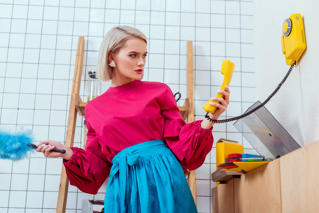 Verwirrte Hausfrau in bunten Klamotten schaut auf Retro-Telefon in Küche - Foto, Bild