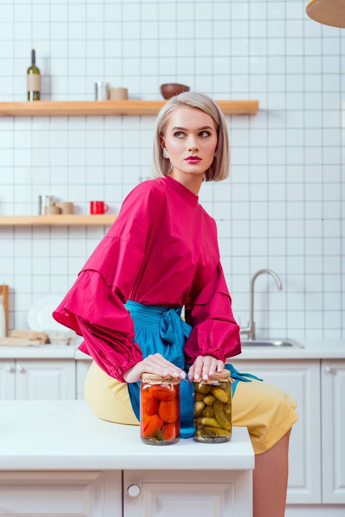 foco seletivo de bela dona de casa na moda sentado no balcão da cozinha com frascos de legumes em conserva na cozinha
  - Foto, Imagem