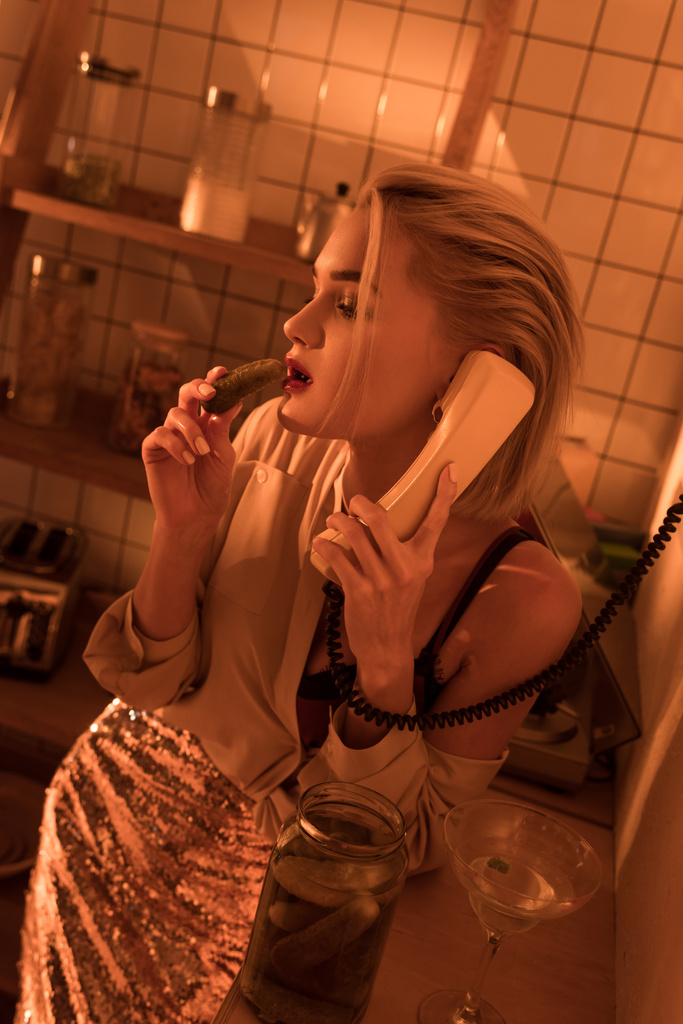 beautiful elegant woman eating pickled cucumber while talking on retro telephone in kitchen with orange light - Photo, Image