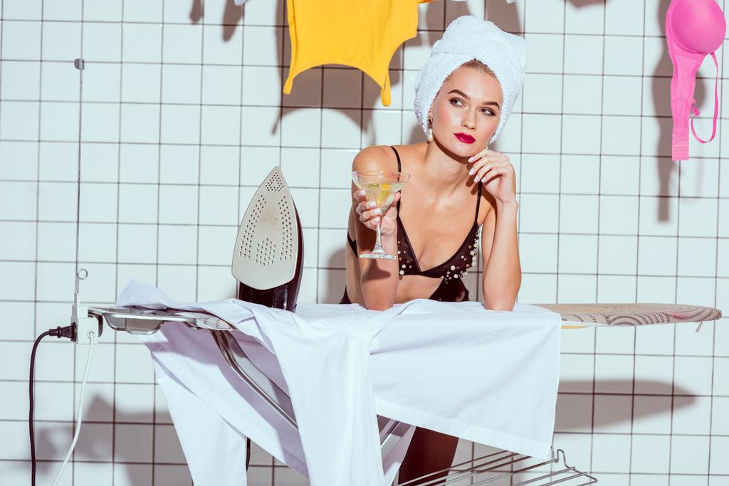 красивая мечтательная домохозяйка в полотенце с коктейльным бокалом во время глажки в ванной комнате
 - Фото, изображение
