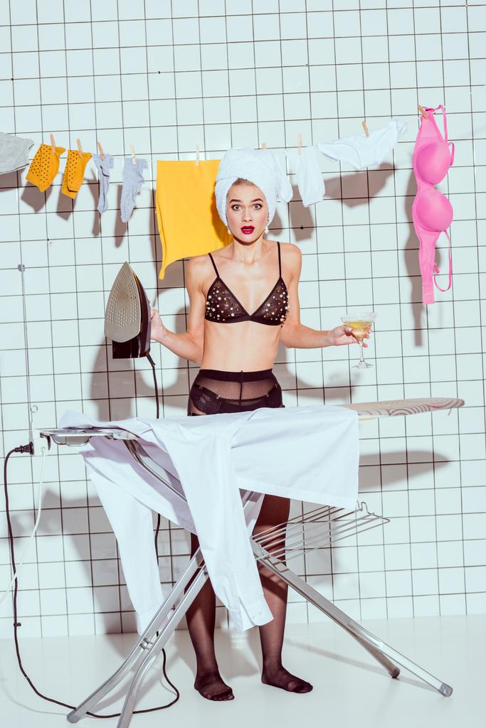 красивая удивленная домохозяйка с коктейлем и полотенцем на голове гладильная одежда в ванной комнате, глядя в камеру
 - Фото, изображение