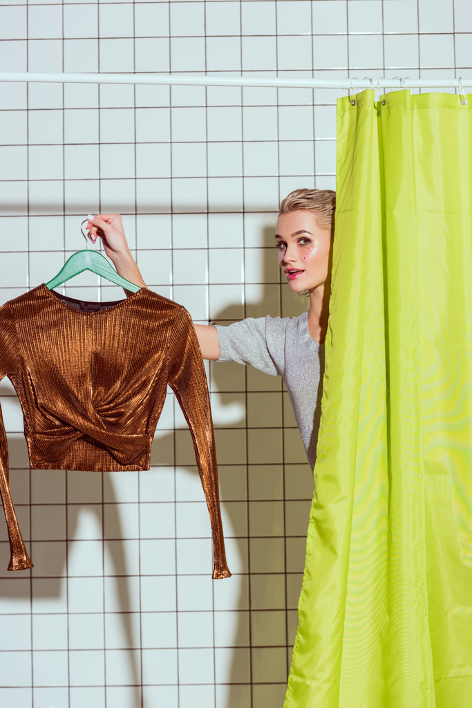 mulher segurando top bronze no cabide e olhando para a câmera no chuveiro com cortina verde
 - Foto, Imagem