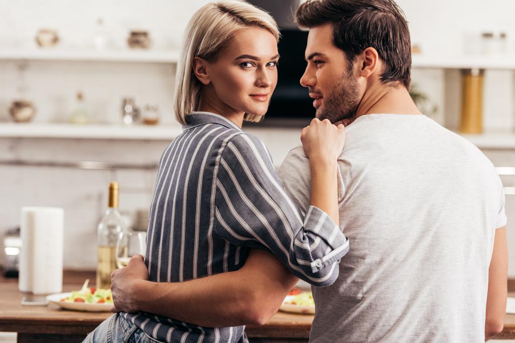 beautiful young couple hugging in kitchen and looking at camera - Photo, Image