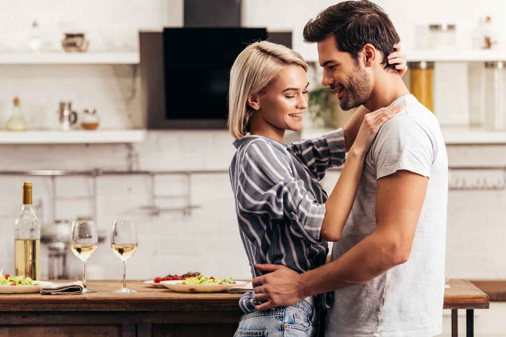 knappe vriendje en aantrekkelijke vriendin glimlachend en knuffelen in keuken - Foto, afbeelding