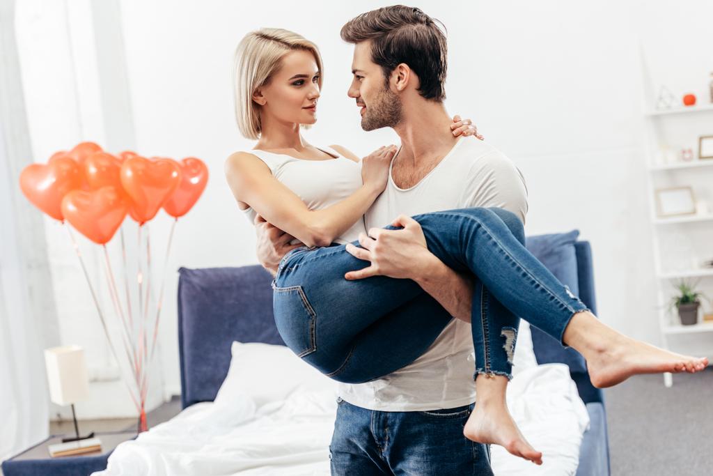 enfoque selectivo de novio guapo sosteniendo novia atractiva en el dormitorio en el día de San Valentín
 - Foto, Imagen
