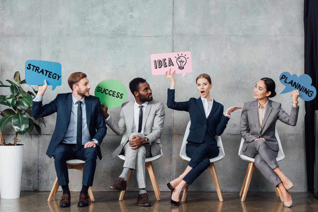 happy multiethnic businesspeople holding speech bubbles with idea, success, planning and strategy lettering while sitting in waiting hall - Photo, Image