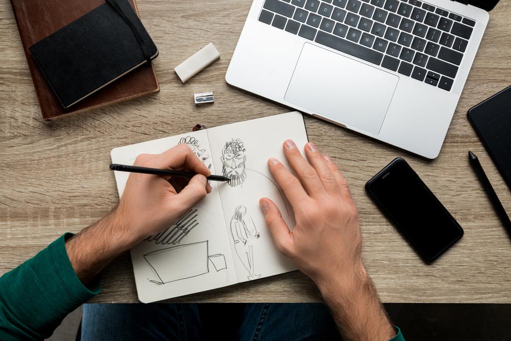 Draufsicht der Hände, die neben dem Laptop auf dem Holztisch auf Album und Smartphone zeichnen - Foto, Bild