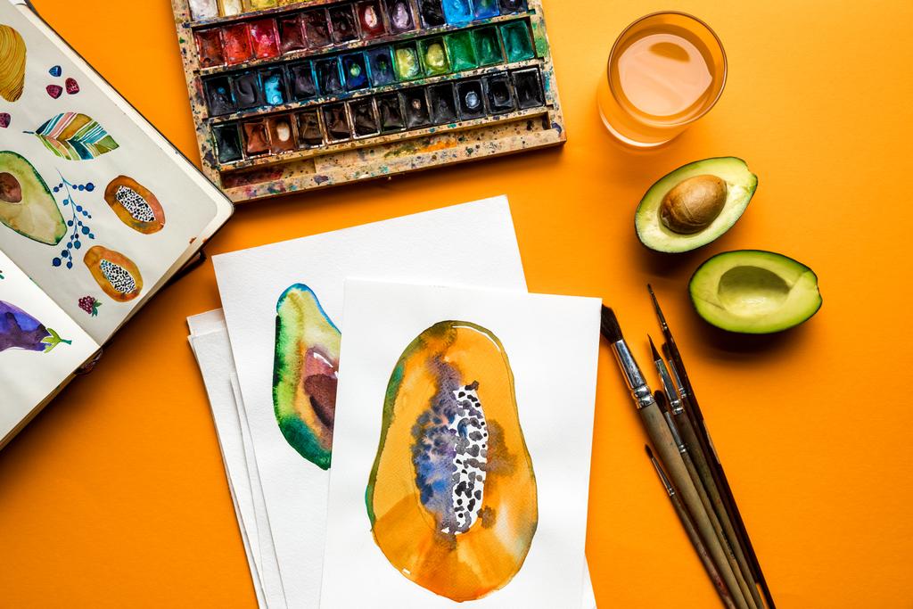 vista superior de tintas coloridas, pincéis, caderno de esboços, desenhos com abacate e mamão sobre fundo amarelo
 - Foto, Imagem