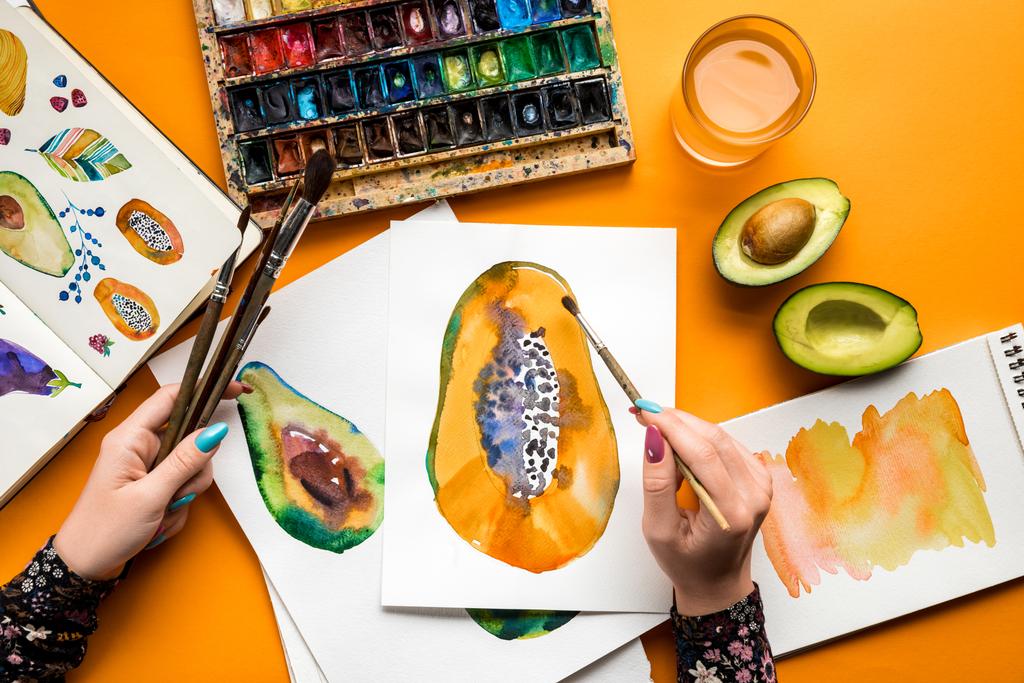 pohled shora ženských rukou, avokádo a papája s akvarel barvy, štětec na žluté tabulky - Fotografie, Obrázek