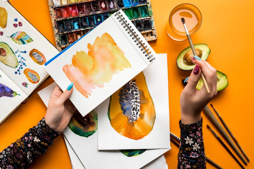 Draufsicht weiblicher Hände, die Avocado und Papaya mit Aquarellfarben zeichnen, Pinsel auf gelbem Tisch - Foto, Bild