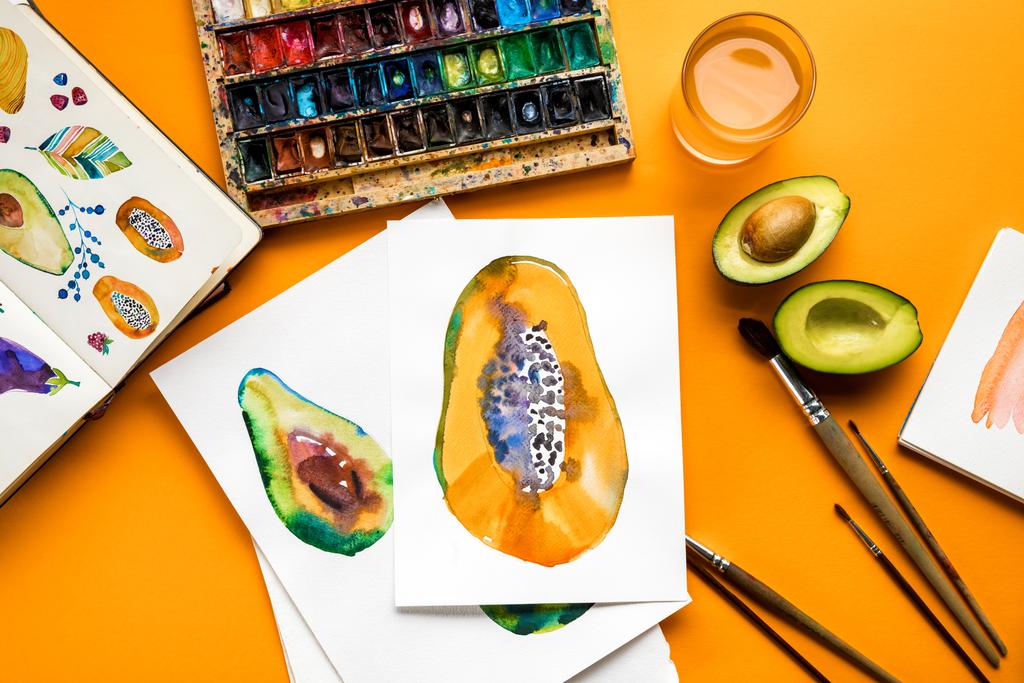 vista superior de los dibujos de aguacate y papaya junto a pinturas de colores, pinceles sobre fondo amarillo
  - Foto, imagen
