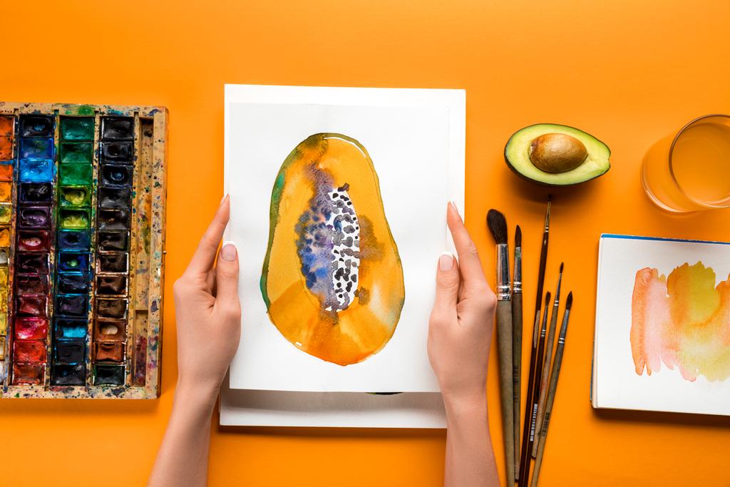 top view of female hands holding papaya picture over yellow table with drawing utensils - Photo, Image