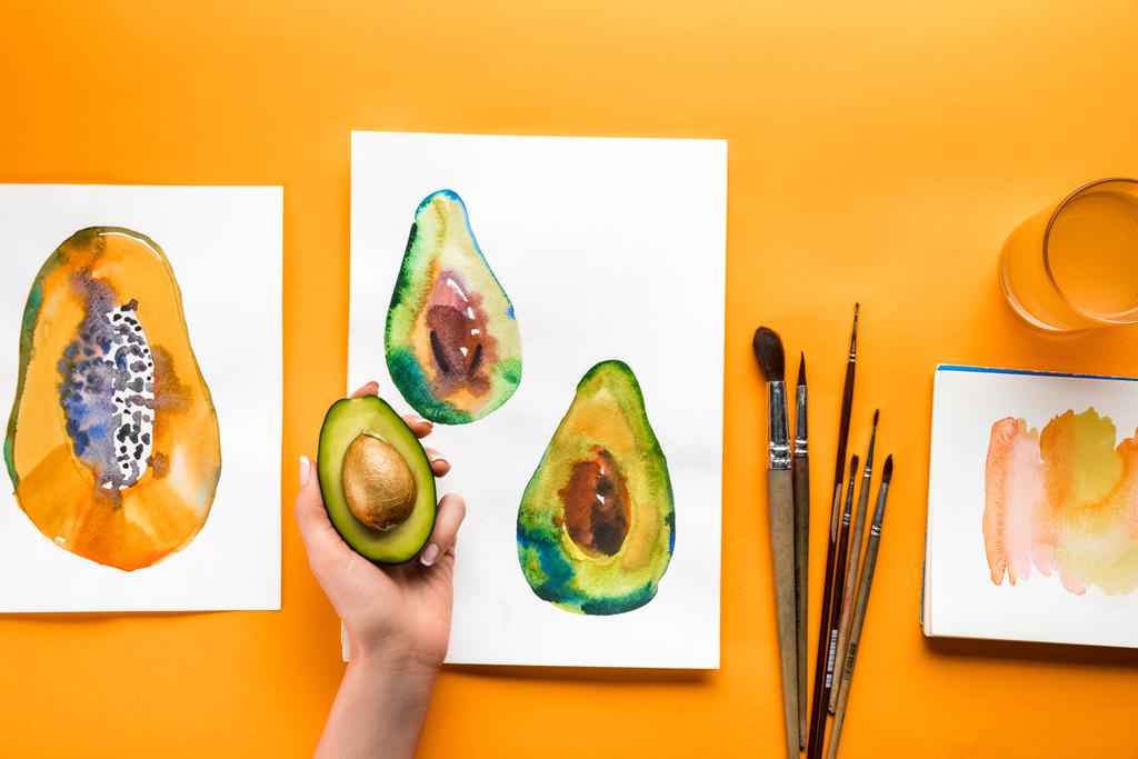 Draufsicht auf weibliche Hand, die Avocado über gelben Tisch hält, mit Zeichnungen von Früchten und Pinseln - Foto, Bild