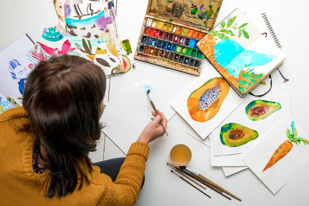 top view of woman painting with watercolors paints while surrounded by color drawings and drawing utensils - Photo, Image