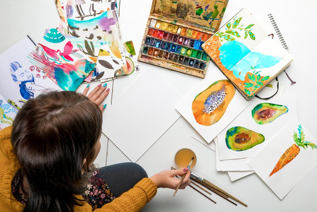 κάτοψη της γυναίκας που ετοιμάζεται να ζωγραφίζει με νερομπογιές χρώματα ενώ περιβάλλεται από τα σχέδια χρώματος και σχεδίασης σκεύη - Φωτογραφία, εικόνα